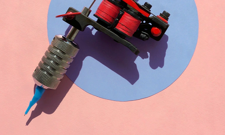A red and black tattoo machine with red spools of thread on a blue circular background over a pink surface.
