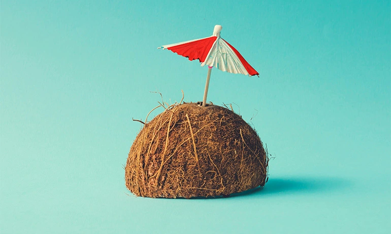 Half of a coconut with a small red and white umbrella sticking out, set against a light blue background.