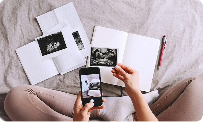 Person holding ultrasound photos while taking a picture with a smartphone, surrounded by open notebooks and more ultrasound images.